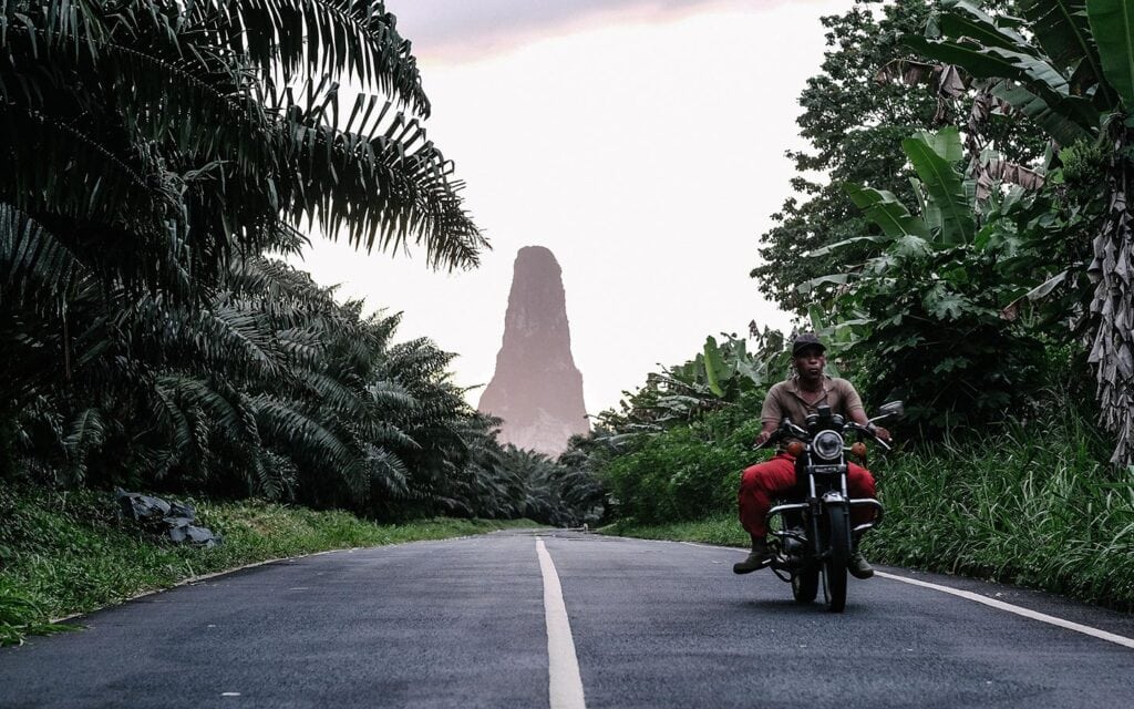 Local man riding a motorbike through southern São Tomé, with Pico Cão Grande rising in the background among rainforest and banana trees.